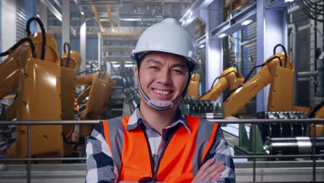 close up of asian male engineer with safety helmet crossing his arms and smiling to camera while standing with industrial robotic arms working