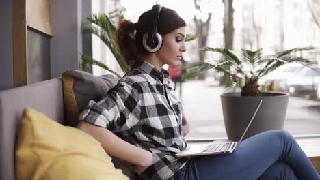 A-girl-with-long-hair-is-sitting-on-the-couch.-She-puts-on-headphones,-adjusts-her-shirt-and-turns-on-her-favorite-music-on-the-laptop.-Side-view.-Flowers-and-window-on-the-background