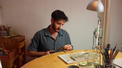 un artesano está sentado en una mesa redonda de madera en un espacio de trabajo brillantemente iluminado, concentrándose en la artesanía detallada, usando herramientas y materiales en su escritorio, rodeado de suministros artísticos.