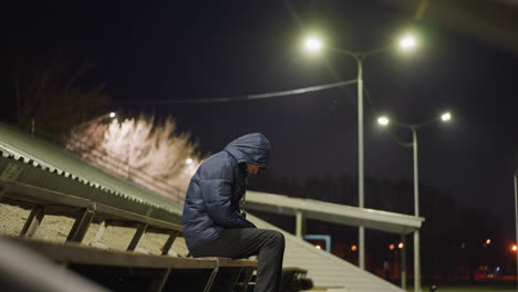 a man wearing a hooded jacket sits alone on bleachers at night, illuminated by streetlights with a dimly lit building and scattered particles in the background