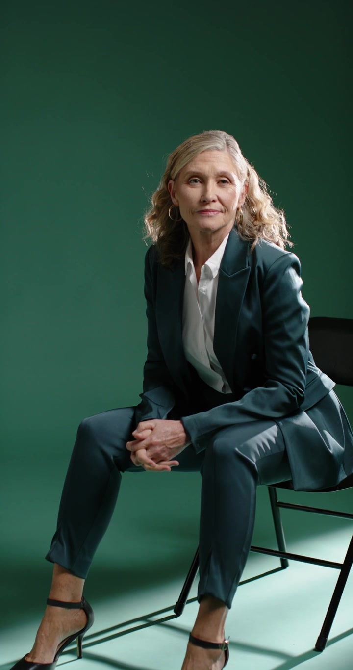 Vertical video: Mature woman sitting on chair in studio in teal suit adjusting hands camera rolling
