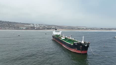 Aerial-panning-shot-of-a-large-black-and-red-ship-freighter-floating-in-the-sea-with-calm-waves-overlooking-the-beach-with-buildings-in-the-background