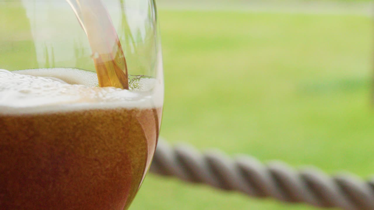 Golden beer pouring into clear glass with foamy head, outdoors in natural daylight, close-up view