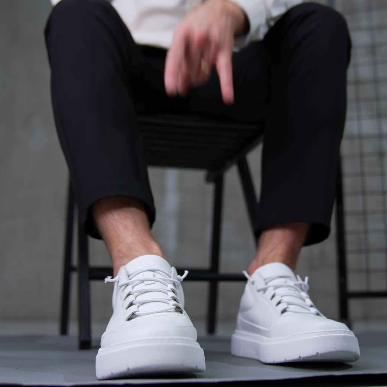 Man wearing black trousers and white shirt presents sneakers. Model sits on chair tapping his foot by the floor. Low angle view