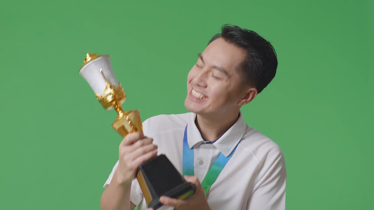 금메달을  아시아인 남자가 황금 트로피를 키스하며, 스튜디오의 녹색 스크린 배경에서 첫 번째 우승자로 자랑스러워합니다.