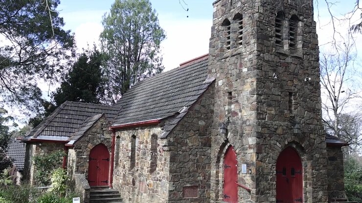 Ampia inquadratura della vecchia chiesa in pietra a Olinda, Melbourne. La muratura invecchiata, le porte rosse e la torre contrastano splendidamente con il fogliame verde sotto una luce diurna soffusa. Pacifico, senza persone.