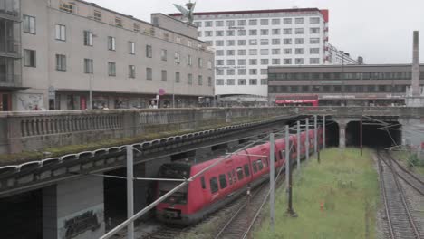 a red passenger train leaving the train station in copenhagen denmark. seen from the bridge as the train goes into the tunnel away from the camera. in the background there are some tall buildings.