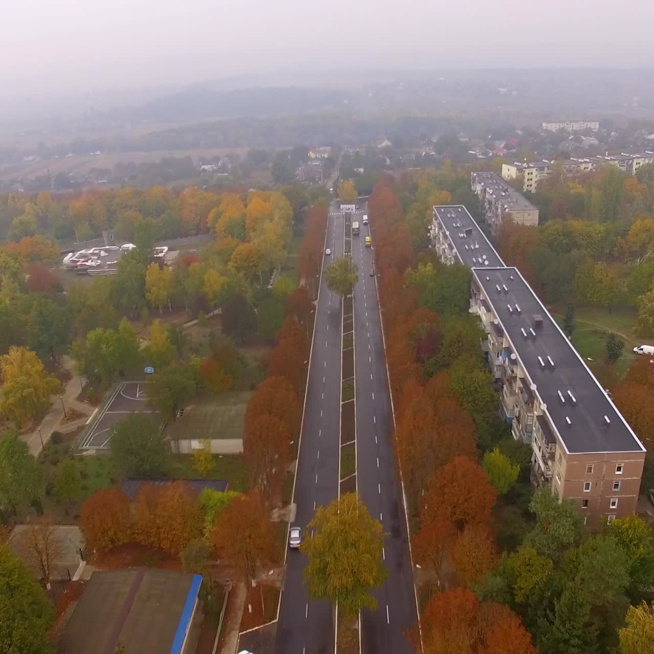 Flying over the road passing through trees colored in green, orange and yellow. Footage over the foggy city residential areas with autumn nature