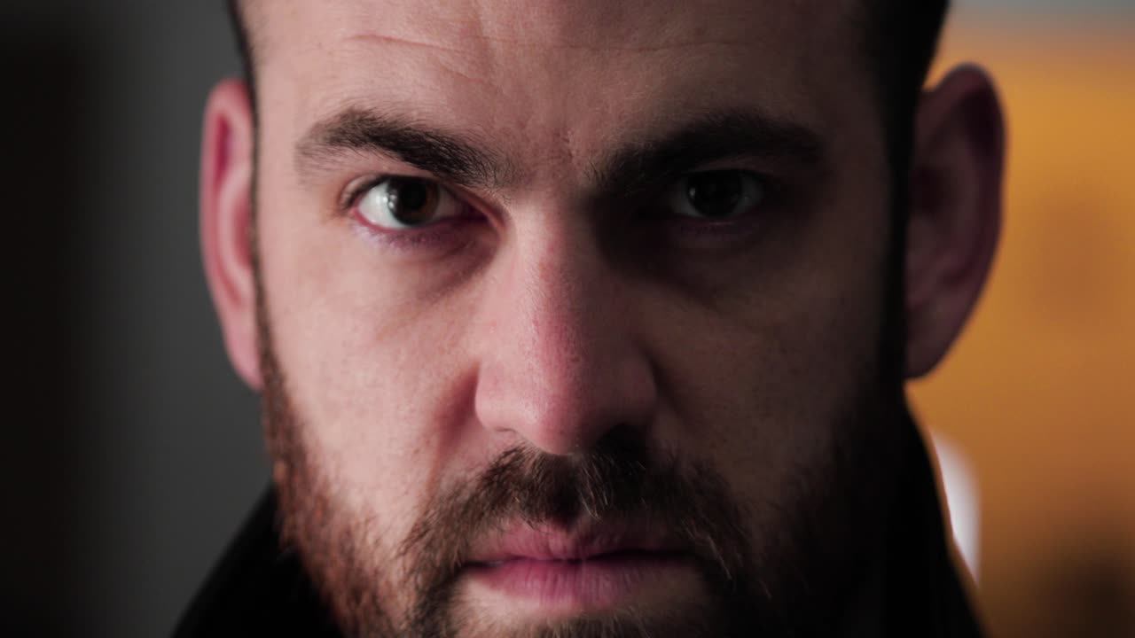 el hombre barbudo mira hacia arriba, temblando de rabia mientras la persona pasa en segundo plano