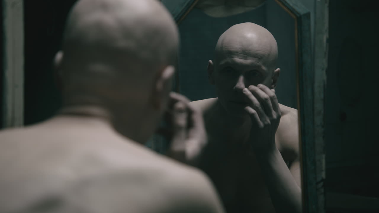 Homem pálido, careca e magro olha-se no espelho no banheiro velho e assustador à noite.