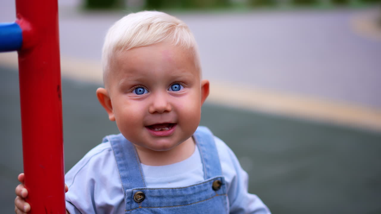 Lovely smiling Caucasian baby stands outdoors holding by the metal ladder. Blond little kid plays at the playground. Blurred backdrop.