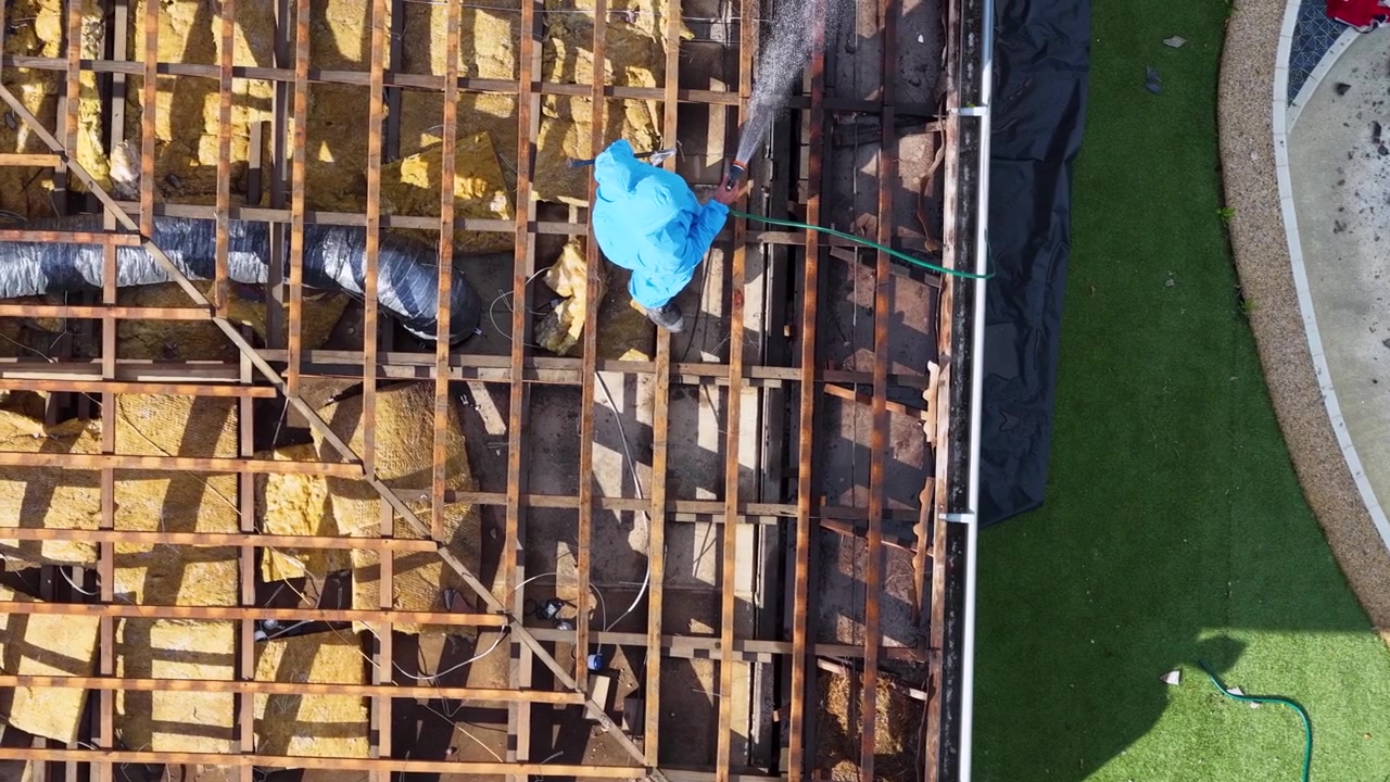 Top-down drone shot of worker in blue protective suit spraying water on wooden roof frame.
