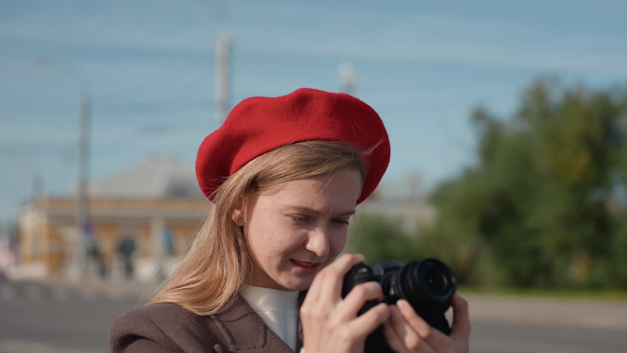 Giovane donna che controlla lo schermo della fotocamera, in una piazza urbana, con basco rosso. Primo piano del controllo di esposizione e messa a fuoco, con un sorriso delicato. Centro commerciale e alberi sullo sfondo, luce dorata. Energia da travel blogger.