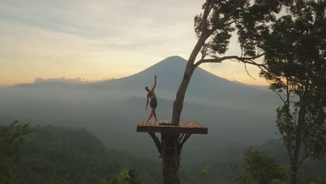 woman practicing standing side stretch yoga pose legs crossed on view platform at mount agung, sunset