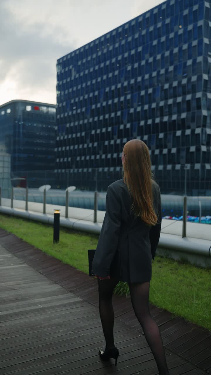 Confident businesswoman with long hair in blazer and heels walks on rooftop terrace surrounded by modern glass buildings