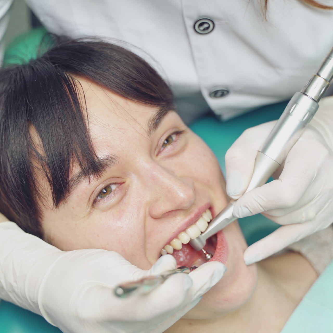 Close-up of patient's face and stomatologist's hands in white gloves at work. Air flow method of cleaning teeth with sandblasting machine in a dental office.