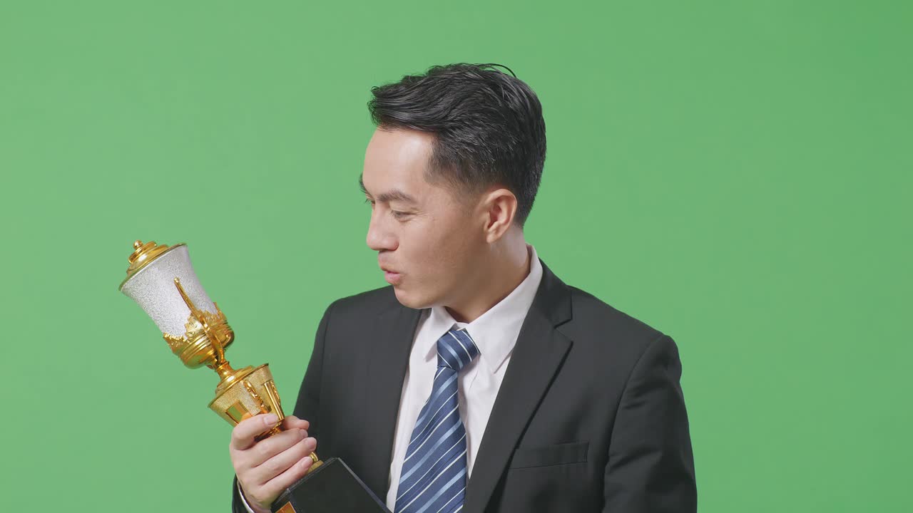 슈트와 타이를 입고 금메달을  아시아 사업가 한 명이 손에 금 트로피를 들고 와우라고 말하며 스튜디오의 녹색 스크린 배경에서 첫 번째 우승자로서 행복합니다.