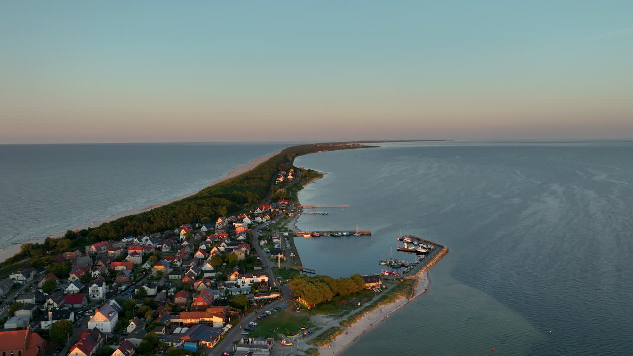Aerial view of Kuźnica, Poland, showing a coastal village with houses, piers, and boats along a narrow strip of land between two bodies of water during sunset