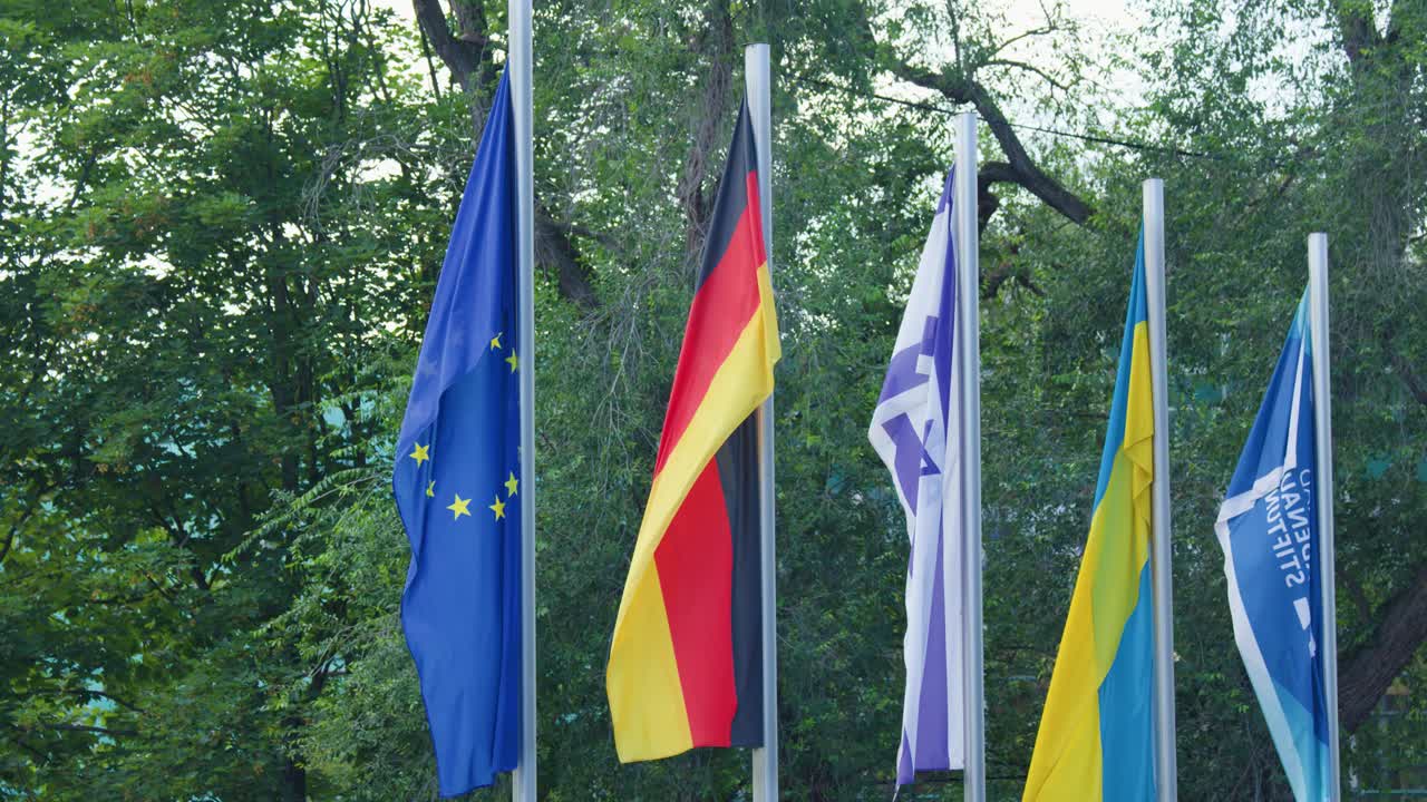 Multiple national and organizational flags wave outdoors, daylight, steady camera, lush green background