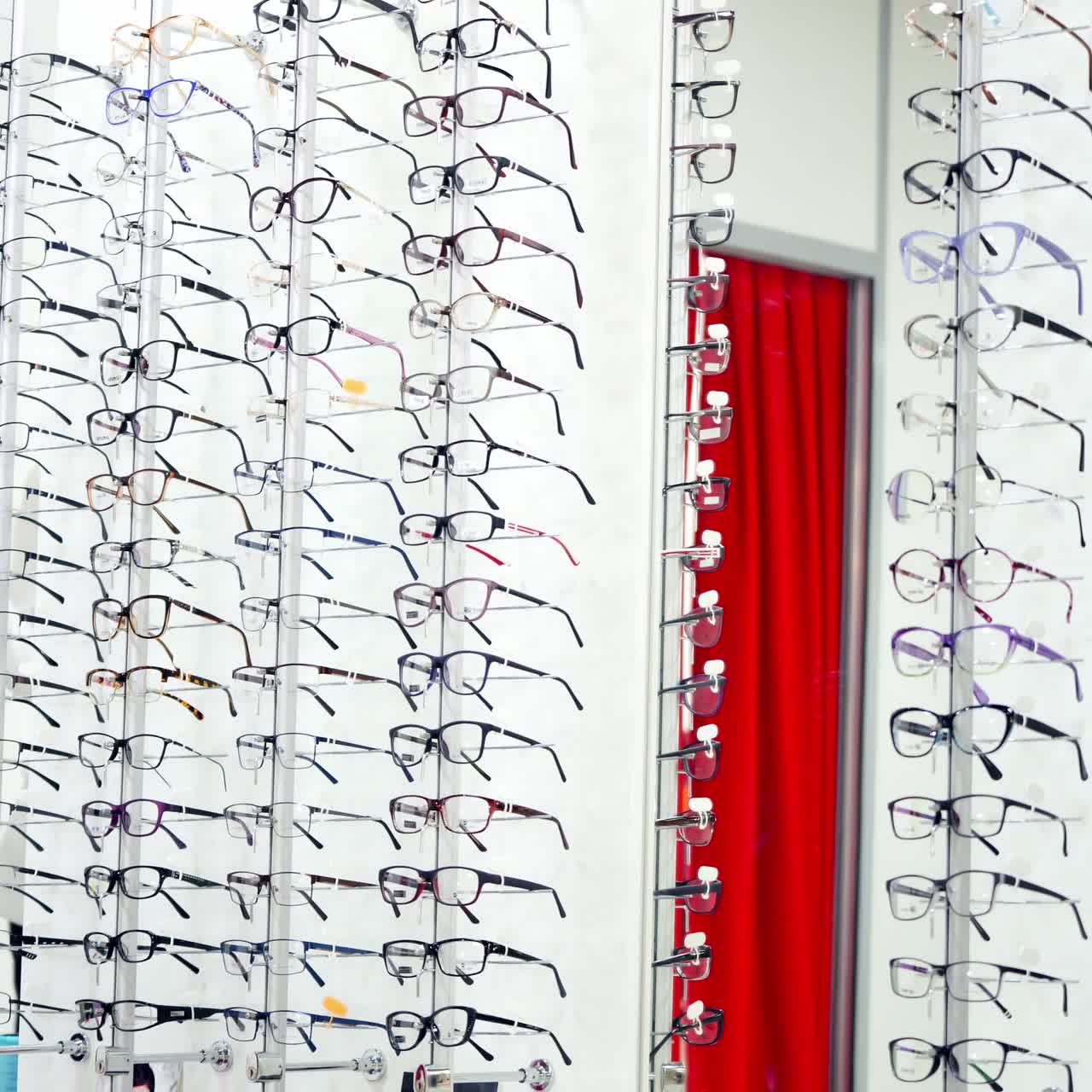 Girl in new glasses looking at camera at optics. Attractive young woman trying on fashionable eyeglasses indoors. Happy buyer choosing spectacles.
