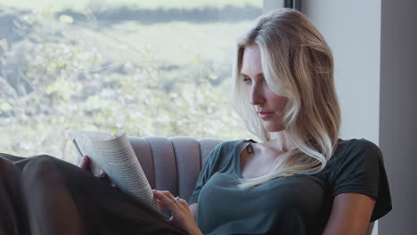 woman relaxing in chair by window at home reading book