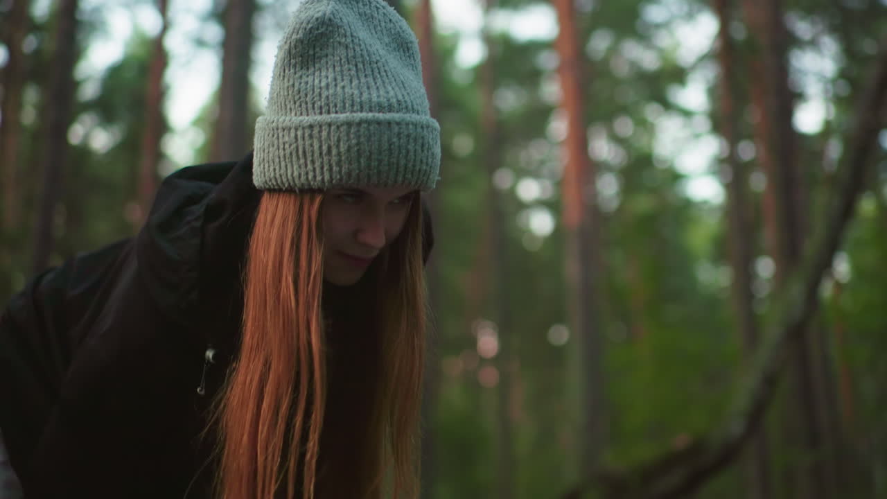Woman in knitted cap and black jacket looks up with focused yet playful expression while engaged in outdoor task in forest, surrounded by tall trees and natural daylight filtering through leaves