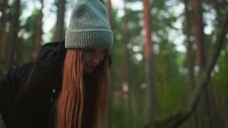 woman in knitted cap and black jacket looks up with focused yet playful expression while engaged in outdoor task in forest, surrounded by tall trees and natural daylight filtering through leaves
