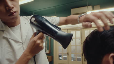 asian hairdresser blow drying hair of client in barbershop