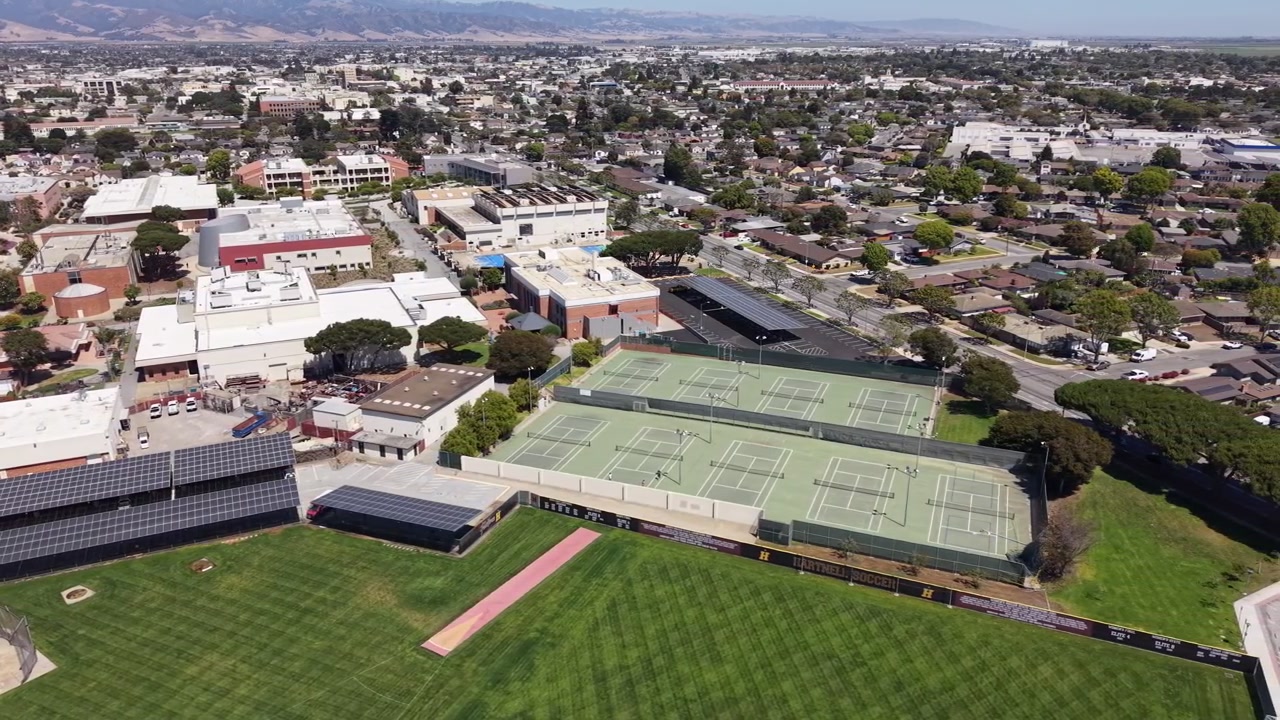カリフォルニア州サリナスにあるハートネル大学の鮮やかな空撮映像。晴れた日に、学術棟、ソーラーパネル、テニスコート、緑のスポーツフィールド、そして広大な市街の景色を捉えています。