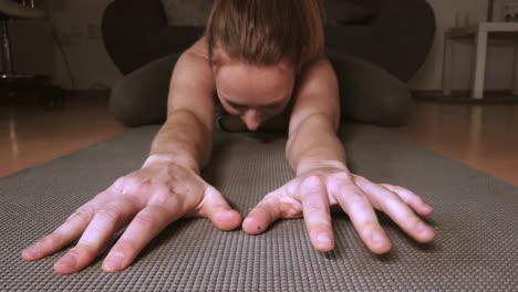 a young woman lowers herself prostrate onto a yoga mat to stretch her arms and fingers toward the camera