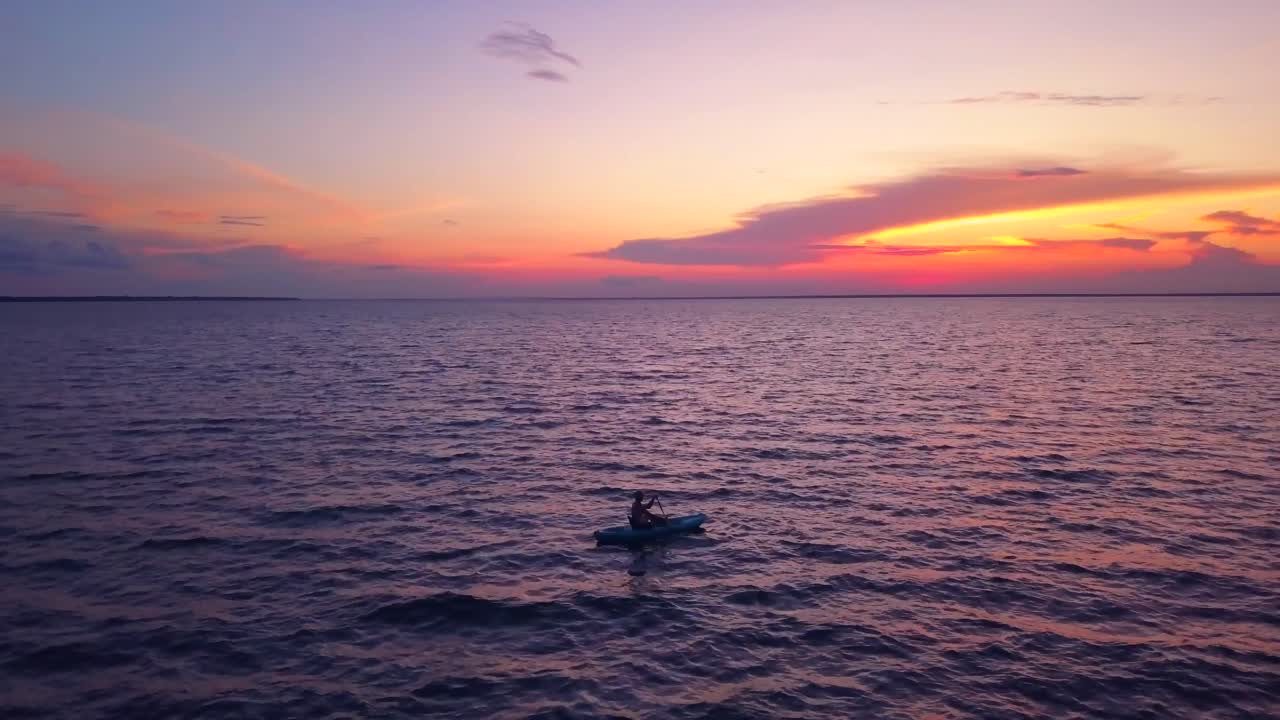 고독한 카약 선수가 형형색색의 일몰 속으로 서서히 흘러가는 극적인 드론 샷