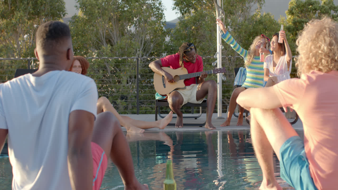 Happy diverse friends with drinks playing guitar at pool party in slow motion
