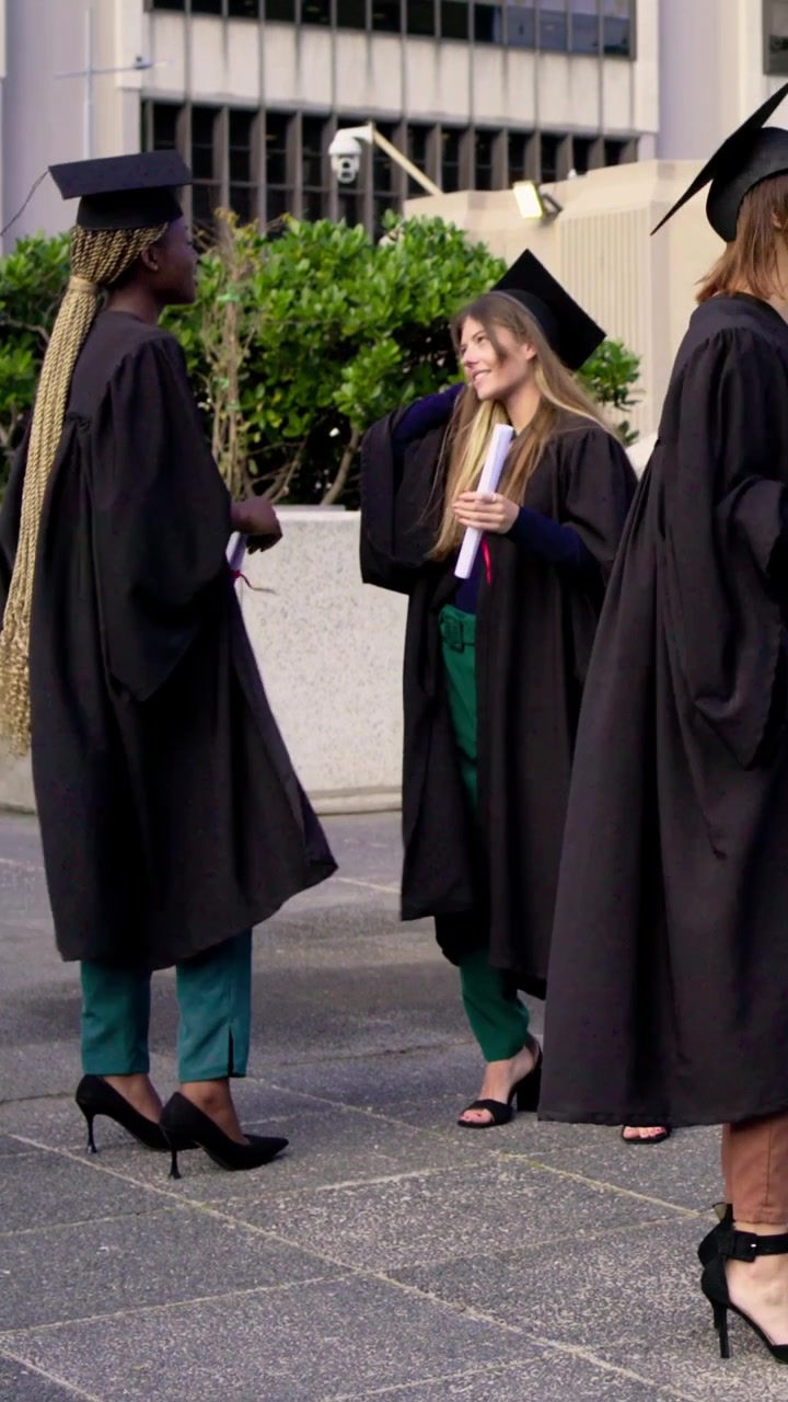 Video verticale: Chiacchierando dopo la cerimonia, due laureati con diplomi in mano in piazza, indossando toghe e tocco.
