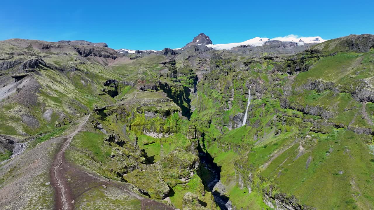 Aerial approaching of iconic Mulagljufur Canyon with waterfall on sunny day with blue sky. Wide shot. Rocky Mountains and green scenery in summer. Iceland, Europe