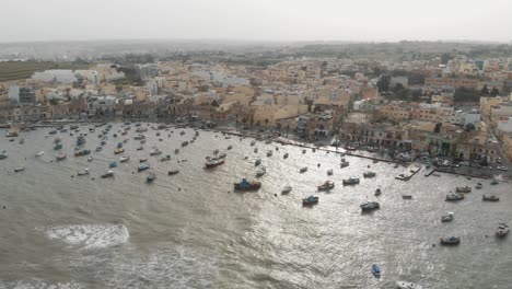 Toma-Panorámica-Aérea-Derecha-Del-Puerto-De-Marsaxlokk-Con-Fondo-De-Pueblo-En-Malta