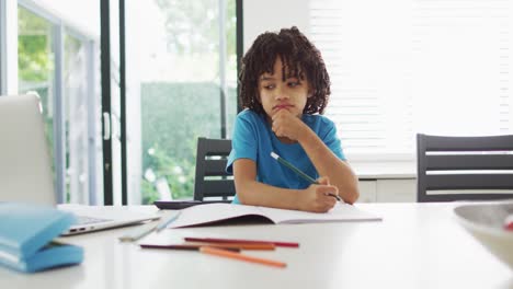 Happy-biracial-boy-sitting-at-table-in-kitchen-doing-homework