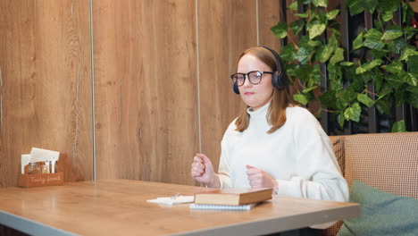 girl with glasses and headphones nods to music with slight dance moves while book rests on wooden table in cozy indoor space with green plants behind displaying relaxed study break vibe