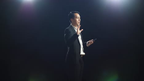 side view of asian speaker man in business suit holding and pointing smartphone while speaking in the black screen studio