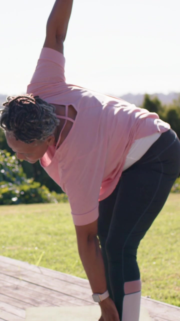 Vertical video: Raising woman cueing friend mirroring reach beside deck, pink tops, mat, watches