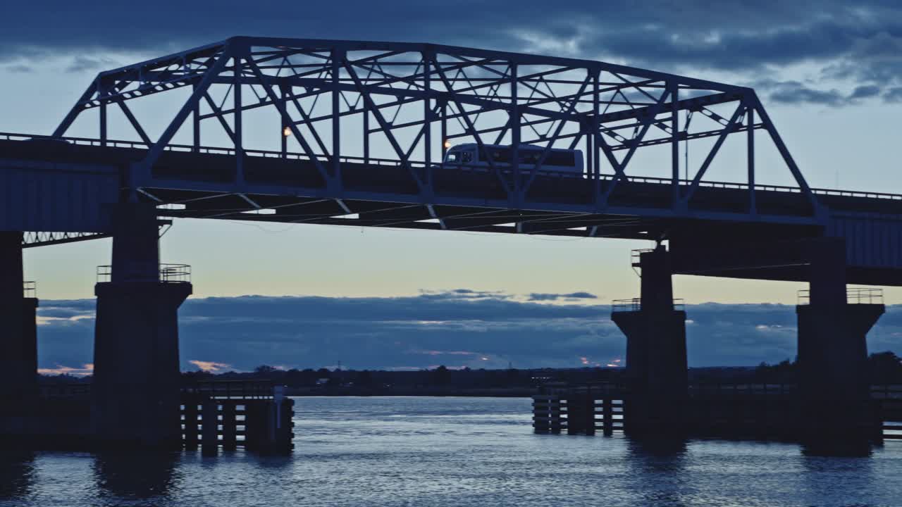 A semi-truck crosses a long bridge over water during a dramatic sunset with moody clouds and soft reflections