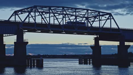 a semi-truck crosses a long bridge over water during a dramatic sunset with moody clouds and soft reflections