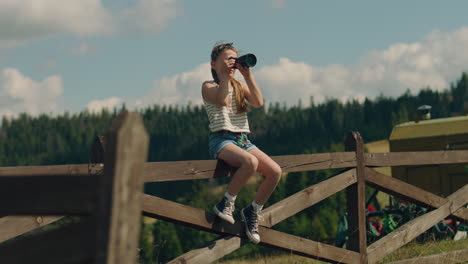 young girl photographer wiping camera lens young photographer wiping digital camera lens by tshirt sitting on wooden fence girl shooting beautiful nature landscape concentrating on process spending leisure time outdoor slow motion