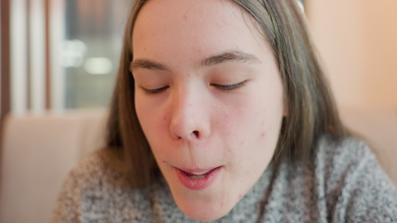 Close up of young girl with long hair licking tea spoon in cafeteria, eyes closed in enjoyment, wearing cozy gray sweater, sitting at table with soft indoor lighting and blurred background atmosphere