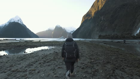 girl in hiking gear walking along shore of milford sound, new zealand