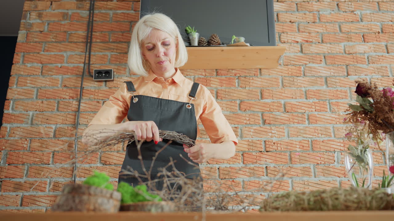Middle aged woman in black apron working indoors arranging dry branches on wooden table with moss and natural materials for rustic decoration project against brick wall background in workshop setting