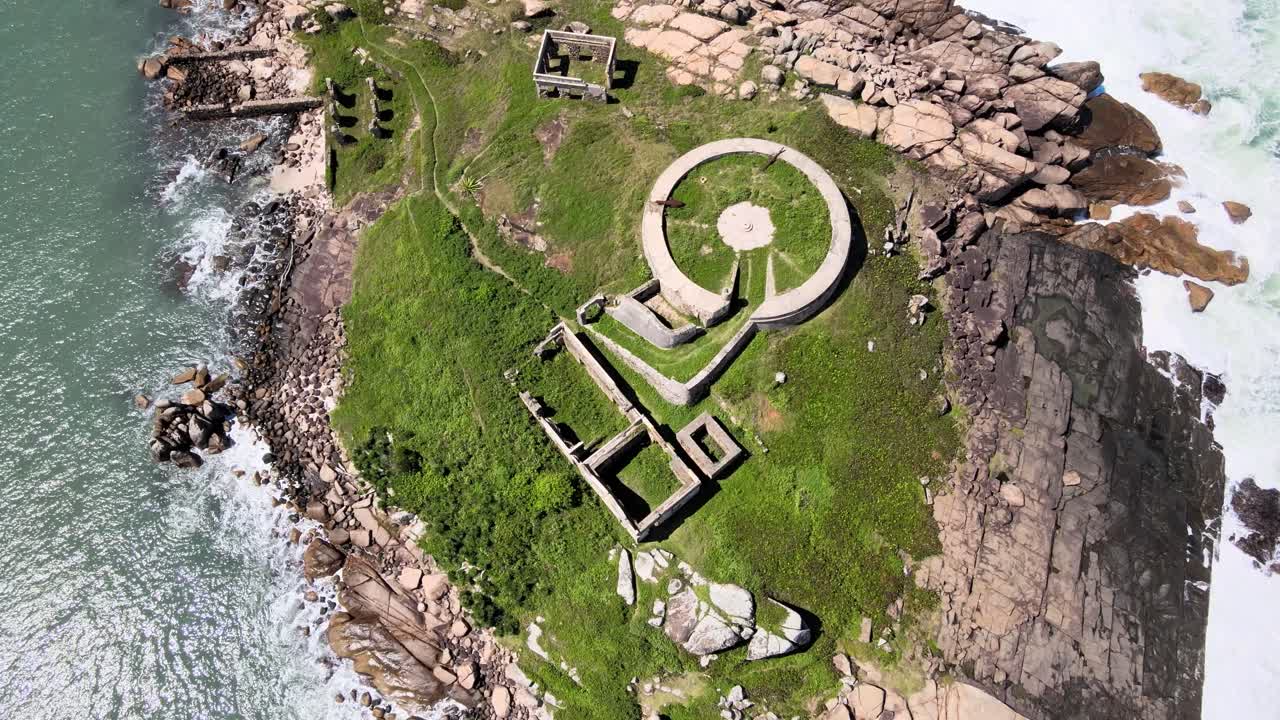 Drone aerial scene of an old abandoned fortress in ruins from the 18th century on an isolated island full of rocks and vegetation surrounded by the sea, Araçatuba Island in Florianópolis