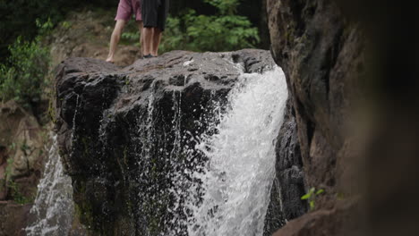 two adventurers stand atop the rugged rock formation at salto el yerba waterfall in misiones, argentina. the cascading water and lush greenery create a lively natural landscape
