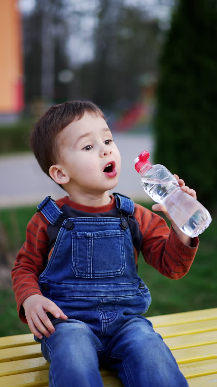 Little cute baby boy sitting outdoors holding a bottle. Kid drinks some water and puts the bottle on the bench. Vertical video.