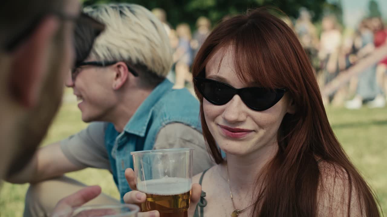 Nærbillede af kaukasisk rødhåret kvinde, der drikker øl fra en engangskop på en musikfestival og snakker med venner.