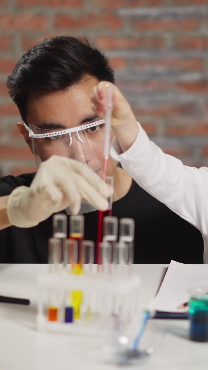 Asian male chemist teaches blonde little child to take liquid with pipette from test tube with researching samples at fun science lesson against brick wall